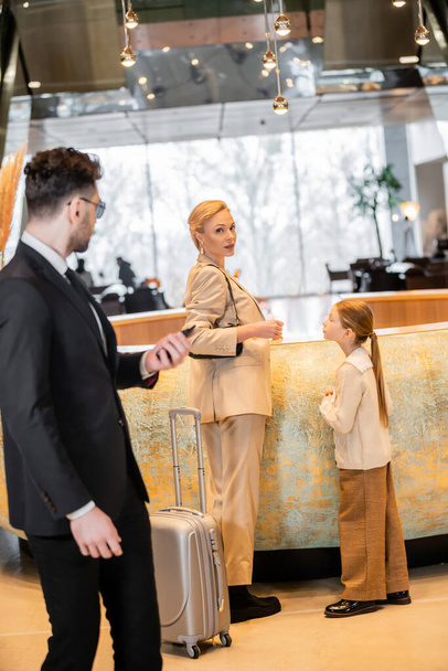 personal security, lifestyle, blonde mother with preteen girl standing at reception desk, successful woman looking at camera during check in, bodyguard in suit standing on blurred foreground  - Foto, imagen
