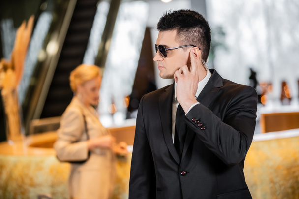 Koruma konsepti, resmi giyimli ve kravatlı yakışıklı bir adam otel lobisinde kulaklığa dokunuyor, güvenlik, iletişim, özel güvenlik, arka plandaki bulanık bayan müşteriyi koruyor.  - Fotoğraf, Görsel