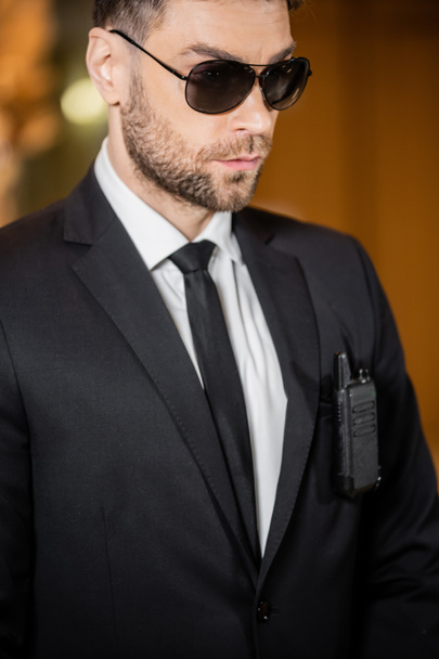 όμορφος σωματοφύλακας, φύλακας σε κοστούμι με γραβάτα και γυαλιά ηλίου στέκεται στο ξενοδοχείο, επαγγελματικές φωτογραφίες κεφαλής, ραδιόφωνο πομποδέκτη που επισυνάπτεται στην τσέπη σακάκι, γενειοφόρος άνθρωπος  - Φωτογραφία, εικόνα