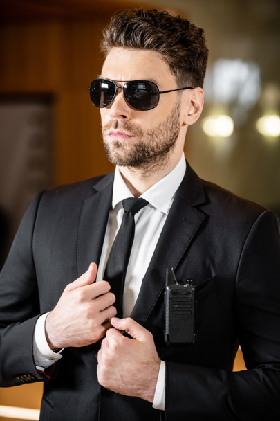 pohledný bodyguard, hlídač v obleku s kravatou a slunečními brýlemi stojící v hotelu, profesionální snímky hlavy, radiový vysílač připevněný k kapse saka, vousatý muž pracující v hotelové ochrance  - Fotografie, Obrázek