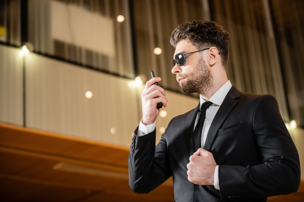 good looking bodyguard, security worker in suit and sunglasses working in lobby of hotel, professional headshots, bearded man using radio transceiver while working in hotel  - Фото, зображення