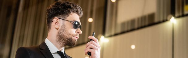 good looking bodyguard, security worker in suit and sunglasses working in lobby of hotel, professional headshots, bearded man using radio transceiver while working in hotel, banner  - 写真・画像