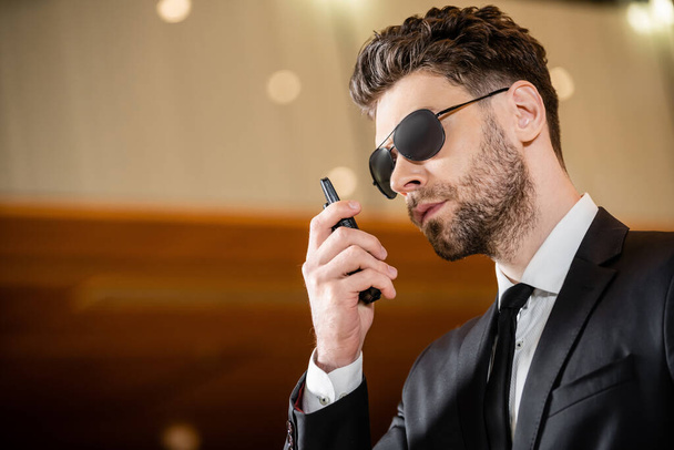 good looking bodyguard, security worker in suit with tie and sunglasses working in lobby of hotel, professional headshots, bearded man using walkie talkie while working in hotel, radio transceiver - Photo, Image