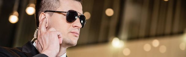 handsome bodyguard in sunglasses, handsome man in suit and tie touching earpiece in lobby of hotel, security, career, communication, vigilance, private safety, hotel staff, banner  - Foto, immagini