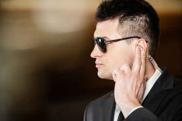 handsome bodyguard in sunglasses, handsome man in suit and tie touching earpiece in lobby of hotel, security, career, communication, vigilance, private safety, hotel work - Foto, Bild