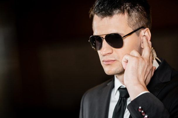 good looking bodyguard in sunglasses, handsome man in suit touching earpiece in lobby of hotel, security, career, communication, vigilance, private safety, hotel security staff, male personnel  - 写真・画像