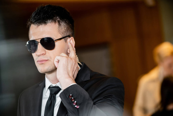 bodyguard v tmavých slunečních brýlích, pohledný muž v obleku a kravatě dotýkající se sluchátka v lobby hotelu, ochranka, kariéra, komunikace, ostražitost, bezpečnost hotelu, mužský personál  - Fotografie, Obrázek