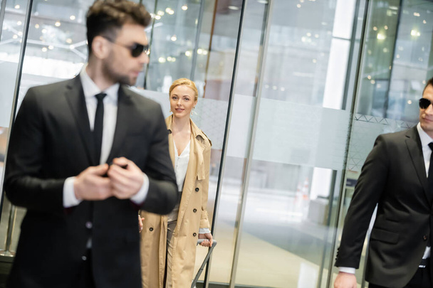 private beveiliging, blonde vrouw in trench jas het hotel binnengaan, wandelen met bagage, twee bodyguards in formele kleding en zonnebril op wazig voorgrond, persoonlijke veiligheid  - Foto, afbeelding