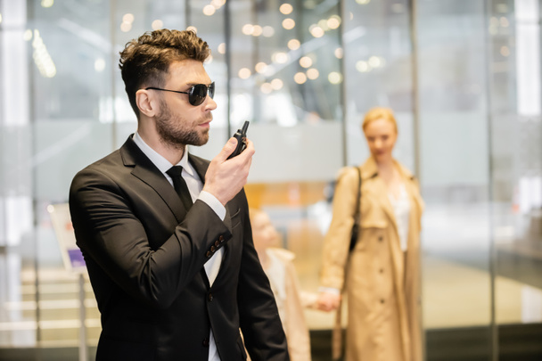 Koruma kavramı, takım elbiseli ve kravatlı yakışıklı adam radyo vericisi kullanıyor, arka planda müşterileri koruyor, telsiz telsizi, bağlantı ve güvenliği elinde tutarken konuşuyor. - Fotoğraf, Görsel