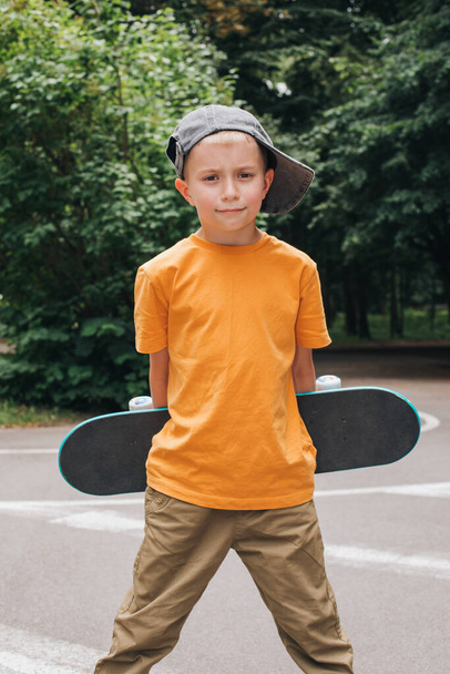 Atividades ao ar livre para crianças. Um menino alegre em um capcstay com um skate em um parque público. Um estudante caucasiano a passear. Vista frontal - Foto, Imagem