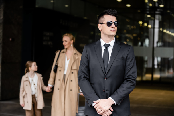 personal security, lifestyle, bodyguard in suit standing near successful woman with preteen child, protecting mother and daughter near hotel, rich life, family travel, private security service  - Photo, Image