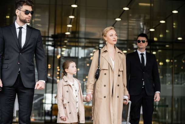 dva bodyguardi kráčející vedle úspěšné ženy a předpuberťačky, vstupující do hotelu, soukromá ochranka, blondýna matka a dcera v pláštích, bezpečnost a ochrana, rodinné cestování, bohatý život  - Fotografie, Obrázek