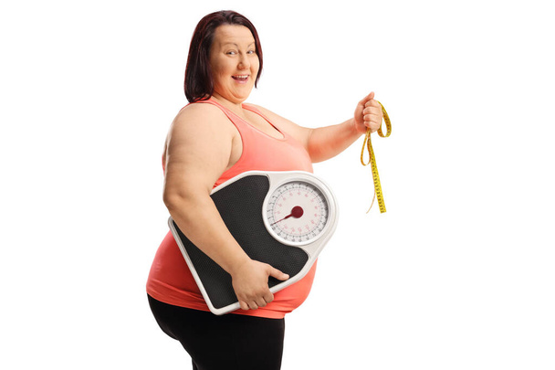 Cheerful overweight woman holding a tape and a weight scale isolated on white background - Фото, изображение