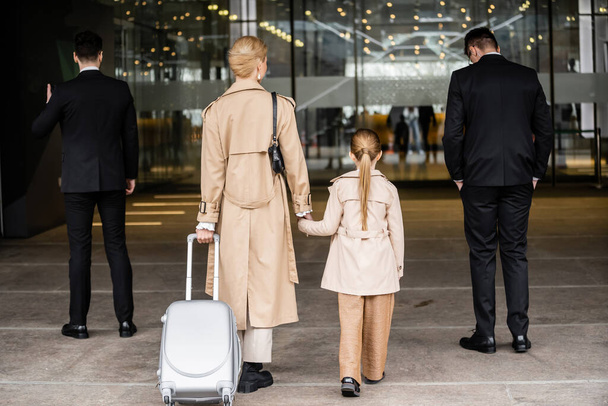 back view of two bodyguards walking next to blonde woman and preteen child, entering hotel, private security, mother holding hands with daughter,  personal safety and protection  - 写真・画像