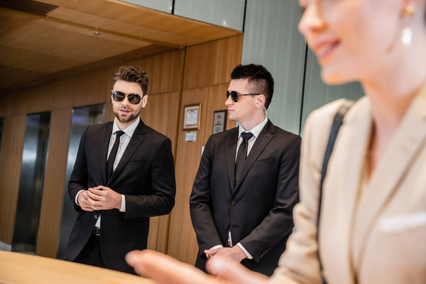 handsome bodyguards protecting successful client in hotel, woman on blurred foreground, security service, personal safety, men in suits and sunglasses, hotel reception  - Photo, Image