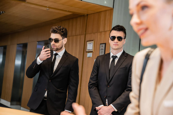 handsome bodyguards protecting beautiful and successful client in hotel, blonde woman on blurred foreground, security service, personal safety, man using walkie talkie  - Photo, Image
