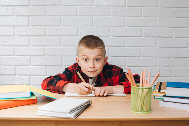 Концепция вернуться в школу. Мальчик в процессе обучения сидит за столом с книгами, блокнотами и карандашами. Вид спереди - Фото, изображение