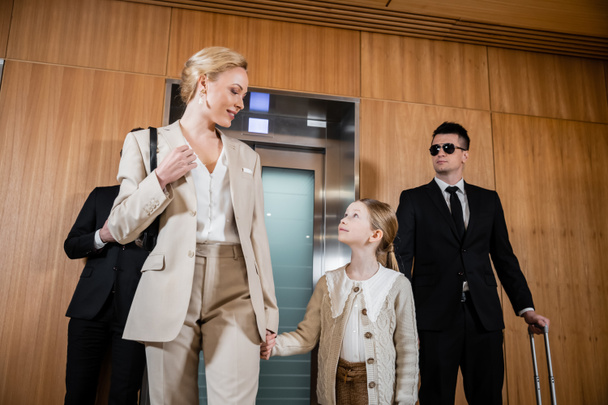 szczęśliwa matka i dziecko trzymając się za ręce stojąc w pobliżu hotelowych wind i ochroniarzy w garniturach, ochrona osobista, kobieta sukcesu i nastoletnia córka, podróże rodzinne, służba bezpieczeństwa  - Zdjęcie, obraz