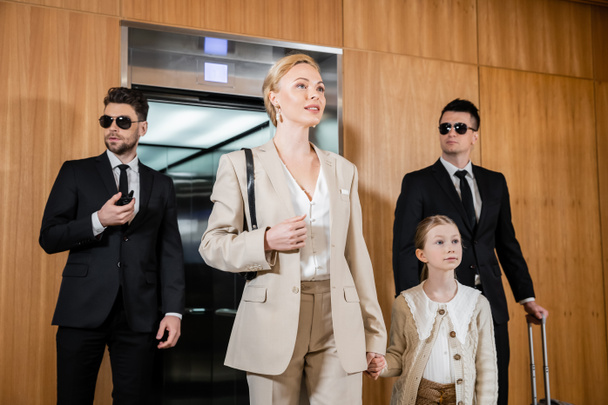 blonde mother and child holding hands while standing near hotel elevator and bodyguards in suits, personal protection, successful woman and preteen daughter, family travel, security service  - Photo, Image