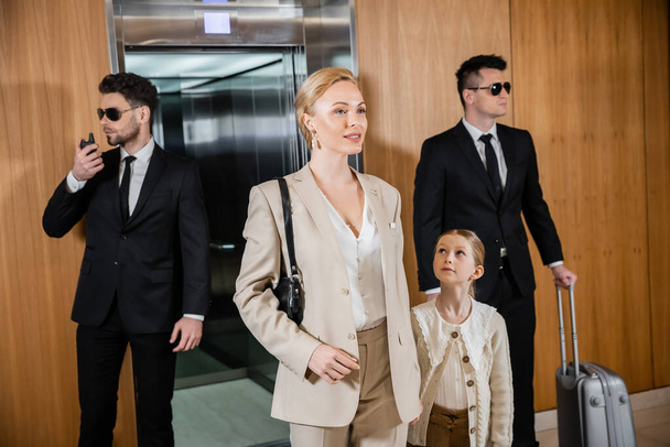 succesvolle moeder en kind hand in hand staan in de buurt van hotel lift en bodyguards in pakken, persoonlijke bescherming, vrouw en tiener dochter, familie reizen, security service  - Foto, afbeelding