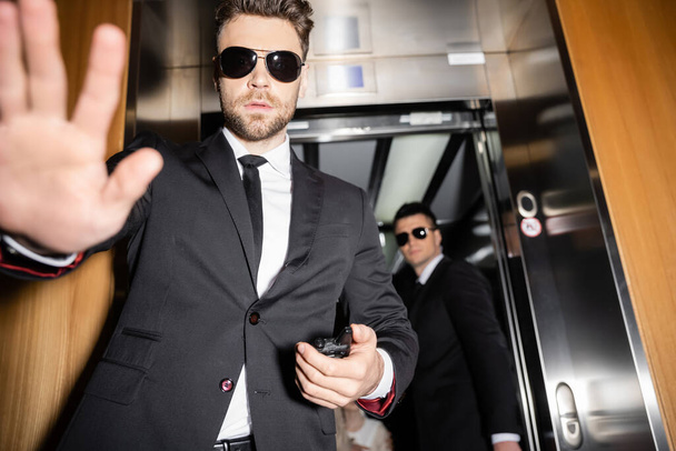 private security concept, handsome bodyguard in sunglasses and suit showing stop, no gesture to camera, protecting private space, hotel lobby, standing next to work partner in elevator  - Photo, Image