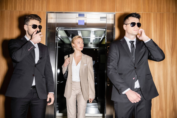 personal security and protection concept, blonde and successful woman with handbag walking out of elevator, bodyguards in suits and sunglasses protecting her privacy in luxury hotel  - Photo, Image