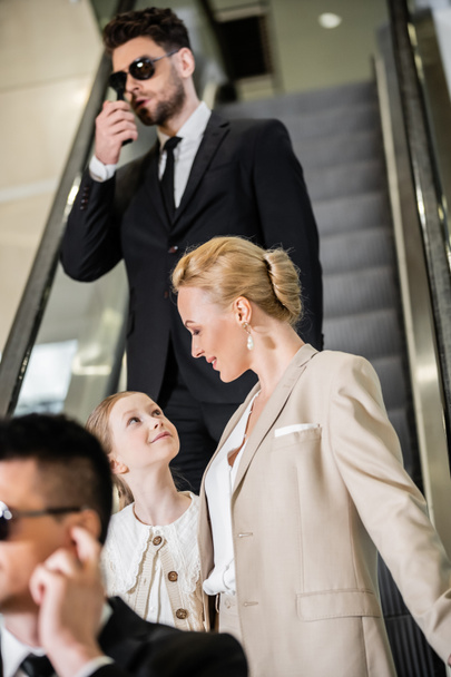 personal security concept, blonde and happy woman and preteen daughter standing on escalator of luxurious hotel, two bodyguards communicating while protecting safety of clients, rich lifestyle  - Photo, Image
