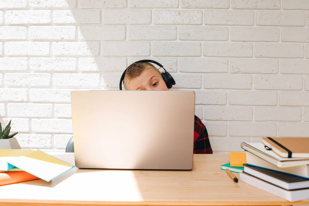 Chłopiec w wieku szkolnym w czarnych słuchawkach siedzi przy stole z laptopem. Dzieci i gadżety. Widok z przodu - Zdjęcie, obraz