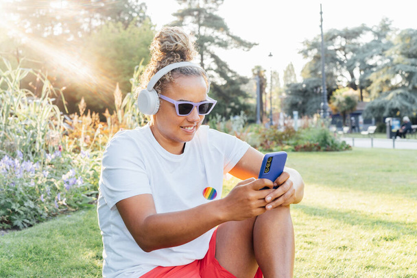 Joven sonriente lesbiana lgbtq mujer utilizando el teléfono móvil en el parque, tener chat de vídeo con amigos, hacer compras en línea, la creación de contenido para los seguidores. Aceptación de la diversidad social, sentirse libre estilo de vida - Foto, Imagen