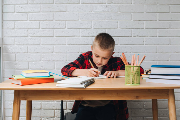 Концепция вернуться в школу. Мальчик в процессе обучения сидит за столом с книгами, блокнотами и карандашами. Вид спереди - Фото, изображение