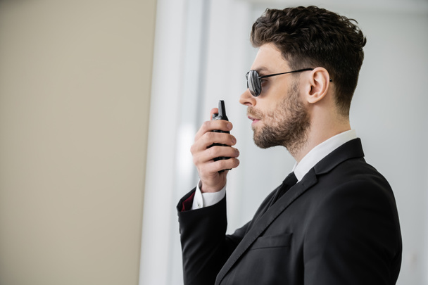 surveillance, bodyguard communicating through walkie talkie, man in sunglasses and black suit with tie, hotel safety, security management, uniformed guard on duty, professional headshots, side view - Photo, Image