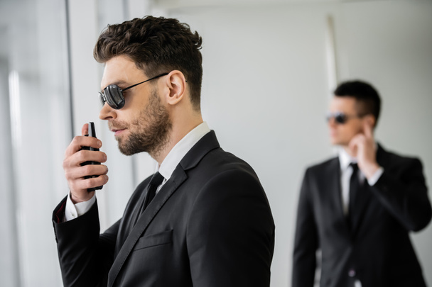 surveillance, bodyguard communicating through walkie talkie, man in sunglasses and black suit with tie, hotel safety, security management, uniformed guard on duty, work partner on background  - Foto, Imagen