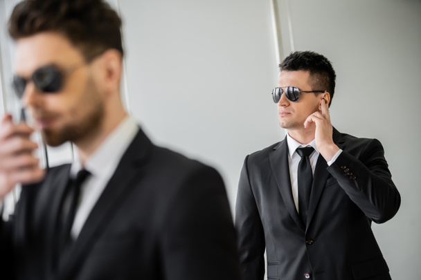 bodyguard communicating through earpiece, man in sunglasses and black suit with tie, hotel safety, security management, surveillance and vigilance, working partner on blurred foreground  - Foto, Imagem