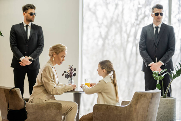 private security concept, bodyguards standing near female clients, preteen girl and blonde mother spending time together in cafe, personal safety and protection, professional in suits  - Photo, Image