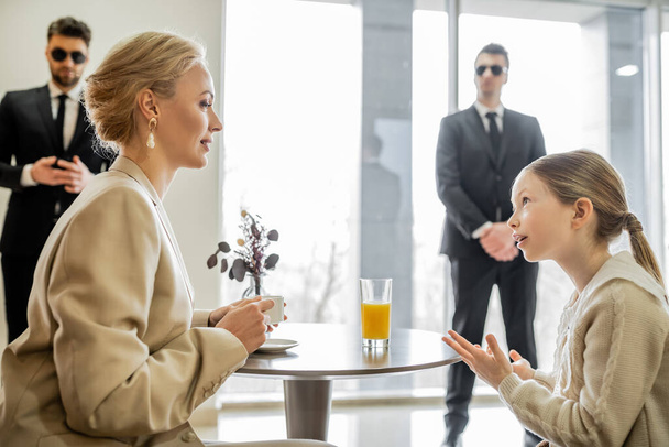 security service, private safety concept, preteen girl talking with blonde mother over drinks in cafe, bodyguards standing on blurred background, personal safety, spending time together  - Photo, Image