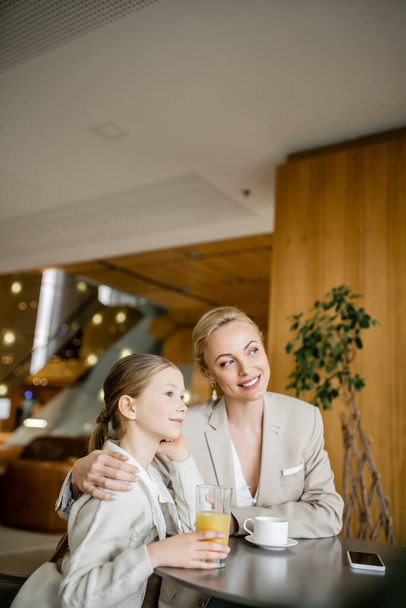 happy mother and daughter spending quality time, blonde woman hugging preteen girl and looking away near beverages and smartphone, working parent and kid, modern parenting, family bonding  - Photo, Image
