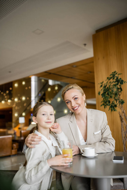 mother and daughter spending quality time together, blonde woman hugging preteen girl and looking at camera near beverages and smartphone, working parent and kid, modern parenting, family bonding  - Zdjęcie, obraz