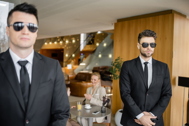 private security service, personal safety concept, handsome bodyguards in formal wear and sunglasses protecting safety of clients on blurred background, working partner, mother and daughter in cafe  - Photo, Image