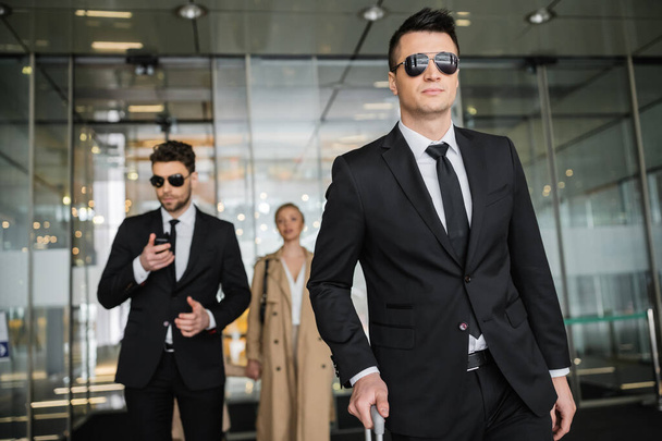 private security service, personal safety concept, handsome bodyguards in formal wear and sunglasses protecting safety of blonde client on blurred background, men and woman walking in hotel  - Photo, Image