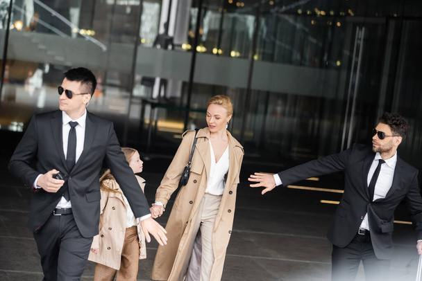 handsome bodyguards protecting safety of female clients, blonde woman and daughter walking out of hotel, safety of rich family, men in sunglasses and suits on duty, work in security  - Foto, immagini
