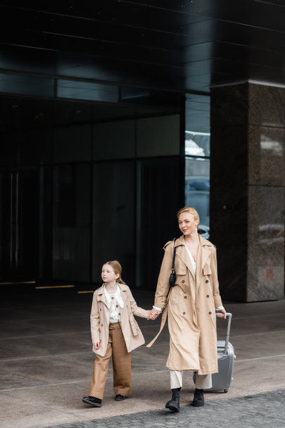 autumn fashion, mother daughter time, happy woman with luggage holding hand of preteen kid while walking out of hotel together, smart casual, beige trench coats, outerwear, trendy look  - Photo, Image