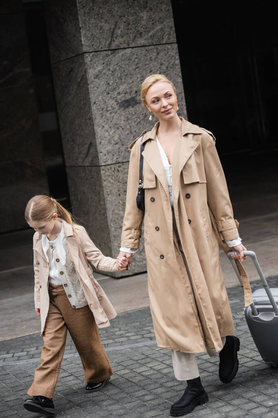 modern ouderschap, moeder dochter tijd, gelukkige vrouw met bagage hand in hand van tiener meisje tijdens het lopen uit hotel samen, slimme casual, beige trench jassen, buitenkleding, trendy look  - Foto, afbeelding