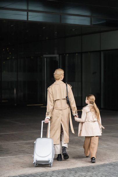 family travel, mother and child, back view of blonde woman with luggage holding hand of girl while walking into hotel together, smart casual, beige trench coats, outerwear, trendy look  - Photo, Image