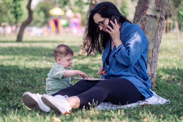Baby boy πατώντας οθόνη tablet και παρεμβαίνουσα μητέρα συνομιλία με τον πελάτη από το κινητό τηλέφωνο κάθεται στο γρασίδι στον κήπο εργασίας ενιαία γυναίκα και ενεργό γιο - Φωτογραφία, εικόνα