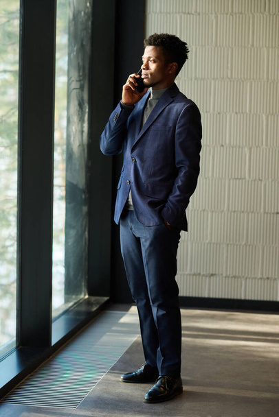 Mladý sebevědomý afroamerický agent v obleku mluví na smartphonu s jedním z klientů, zatímco se dívá přes velké okno v chodbě - Fotografie, Obrázek