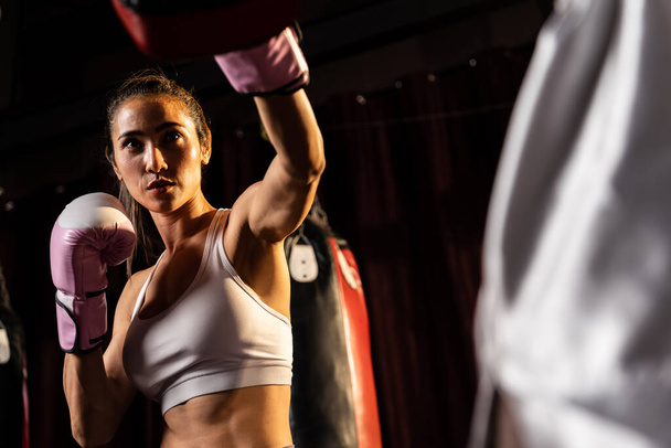 Asian female Muay Thai boxer punching in fierce boxing training session, delivering strike to her sparring trainer wearing punching mitts, showcasing Muay Thai boxing technique and skill. Impetus - 写真・画像