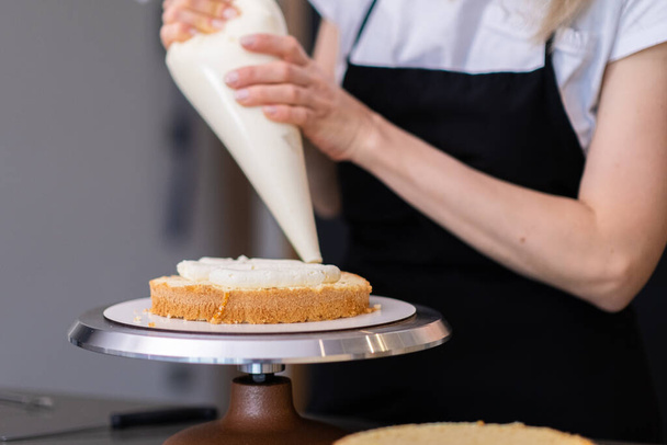 Skilled housewife putting sweet cream on fresh cake layers with big pastry cone woman in black apron standing at kitchen table creating custom confectionery for wedding party closeup - Photo, Image