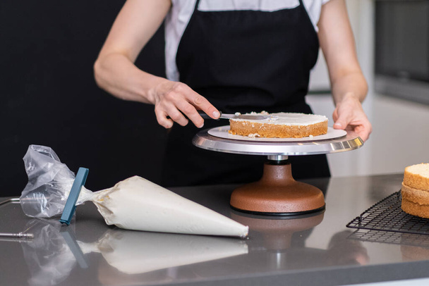 Kobieta w czarnym fartuchu smarowanie organiczne bezcukrowe warstwy ciasta z pysznym kremem za pomocą naczynia kuchennego cukiernik stojący przy stole podejmowania niestandardowe ciasto na uroczystości ślubne - Zdjęcie, obraz
