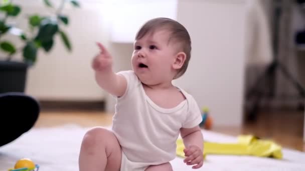 Beyaz elbiseli tombul bir çocuk bembeyaz bir halının üzerine oturmuş bebek odasında çıplak ayakla el hareketleriyle işaret ediyor. Mutlu bir çocukluk ve gelişim dönemi geçiriyor. - Video, Çekim