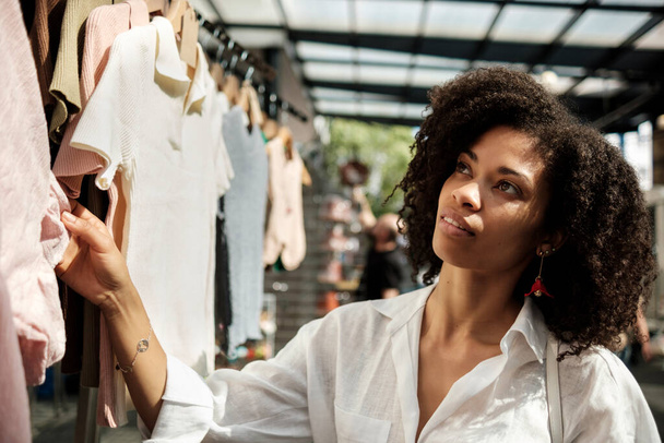 Black woman looking outfits in street market. There are many items hanging on hangers. The market is under roof but model is lit by sunlight. She is wearing a fresh white shirt and some jewelry. Traditional shopping concept. - Foto, Imagem